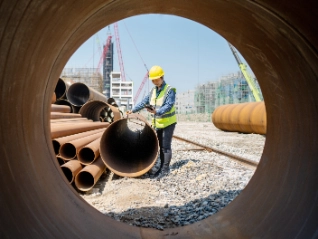 Engineer/Worker (yellow hard hat/vest) holds tablet, inspecting inside of a large metal pipe on a construction site, framed by a circular pipe opening