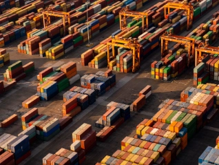 Aerial view: Hundreds of brightly colored shipping containers are stacked high at a busy port/terminal, with large yellow gantry cranes overhead.